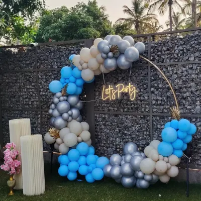 A chic and modern "Let's Party" setup perfect for any celebration. This outdoor decor features a circular frame with a blue, silver, and white balloon garland and a neon sign, creating a stylish photo spot.