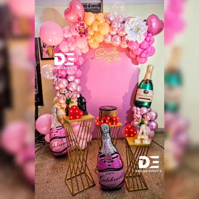 A fun and feminine "Bride to be" decor setup for a bridal shower. The pink backdrop is framed by a balloon arch, and the scene is accessorized with champagne bottle balloons and modern gold stands.
