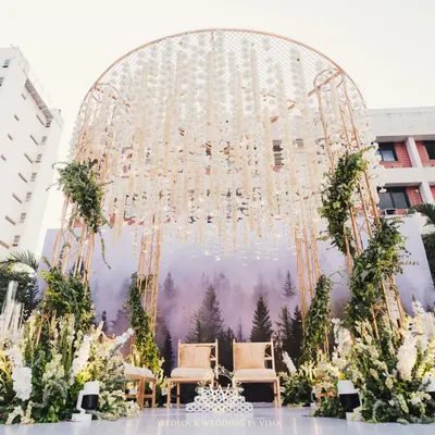 A modern minimalist mandap that perfectly blends nature and contemporary design. The structure features a backdrop of a misty forest, a canopy of hanging white florals, and lush green arrangements.