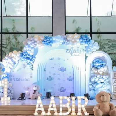 A full view of a dreamy blue and white teddy bear first birthday stage. The layered arches, cascading balloons, and large marquee letters create a stunning focal point for photos and celebrations.