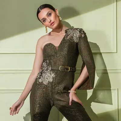 This unique olive green jumpsuit is crafted from a textured, brocade-like fabric. It features a one-shoulder design with a cape-sleeve and is cinched at the waist with a belt for a flattering, modern look.