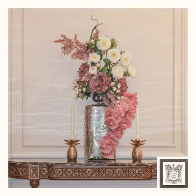 This console is styled with blooming elegance, featuring a dramatic floral arrangement in a tall silver vase. The display is flanked by two pineapple-shaped candle holders, adding a touch of tropical, traditional glow.