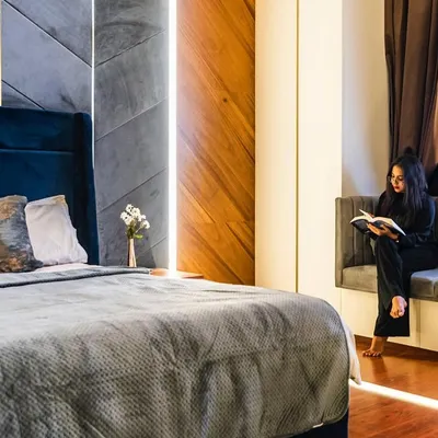 This master bedroom is designed as a cozy retreat. A comfortable reading nook is built into the bay window, and the deep blue velvet headboard is flanked by a geometric-paneled wall with brass inlays. The rich wood flooring and layered lighting create a warm, enveloping ambiance.