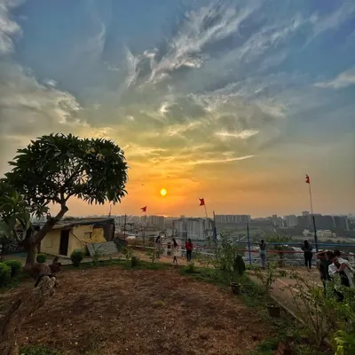 A beautiful sunset view from a hilltop in Mumbai. The image captures people enjoying the view, with the sun dipping towards the horizon over the distant city, creating a serene and picturesque scene.