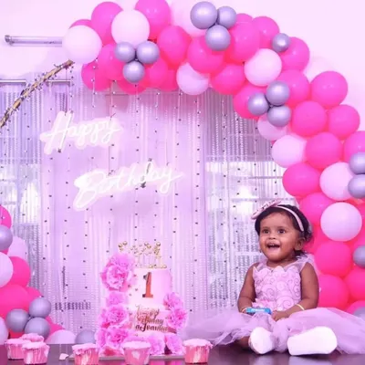 A precious moment from a first birthday party, featuring a beautiful pink and white balloon arch against a shimmer curtain backdrop. The setup includes a custom cake and a "Happy Birthday" neon sign.