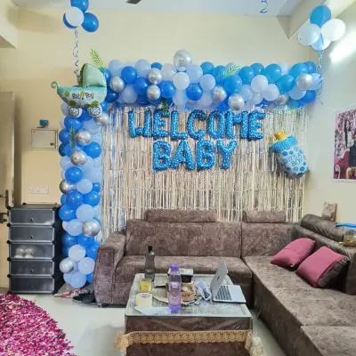 A 'Welcome Baby' decoration for a baby boy in a living room. The setup includes a blue and white balloon frame, a silver shimmer curtain, and baby-themed foil balloons.