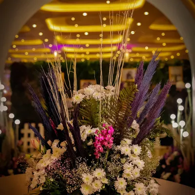 A custom floral centerpiece designed for an evening event. This arrangement combines white chrysanthemums and baby's breath with pops of pink and purple, adding texture and color against a backdrop of warm ambient lighting.