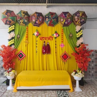 A traditional Haldi stage featuring a bright yellow backdrop, a line of decorative Rajasthani umbrellas, and hanging puppets. The setup is framed with marigold garlands and accented with colorful artificial foliage for a full, festive look.