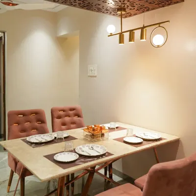 A cozy and elegant dining area. The space features a marble-top table with rose gold legs, comfortable upholstered chairs, and a decorative wooden ceiling panel with a modern chandelier.