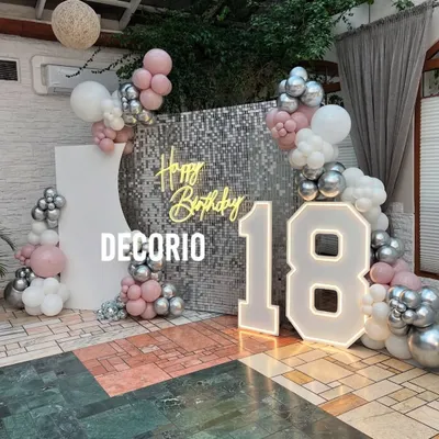 A glamorous 18th birthday setup featuring a silver shimmer wall, a 'Happy Birthday' neon sign, and large marquee numbers. The pink, white, and silver balloon arrangement adds a soft and celebratory touch.