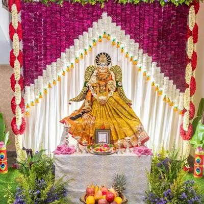 An elegant decoration for the Varalakshmi festival. The goddess is placed before a beautiful curtain of purple and white flowers, creating a divine and colorful backdrop for the pooja.
