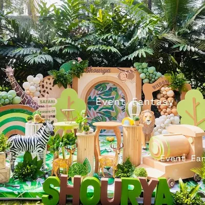 A complete safari adventure setup for Shourya's first birthday. This wide-angle view showcases a lush green outdoor space transformed with a large backdrop, a variety of animal props including a gorilla and zebra, and a custom name display.