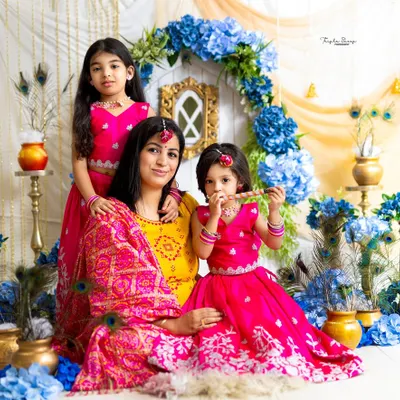 A beautiful Janmashtami-themed portrait of a mother and her two daughters dressed as little Radhas. The vibrant pinks and blues of the set create a festive feel.