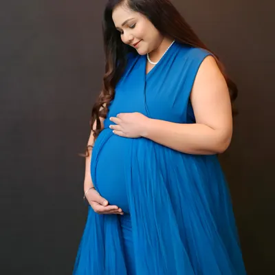 A serene solo portrait of an expecting mother in a beautiful blue gown. This studio shot uses a simple, dark backdrop to emphasize her radiant glow and the gentle embrace of her baby bump.