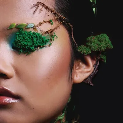 A close-up from the Elle India 'Becoming Earth' editorial, focusing on the intricate makeup detail. The use of moss, twigs, and textured green pigment transforms the face into a natural landscape.
