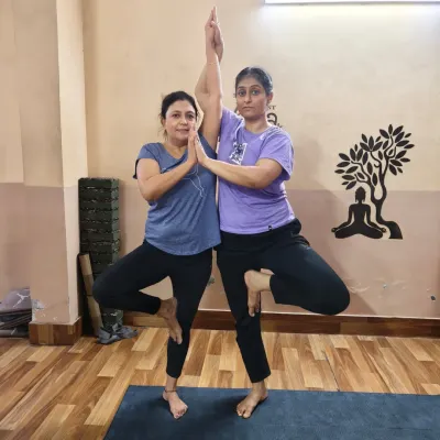 Two students practicing a partner variation of Vrikshasana (Tree Pose). This pose challenges individual balance while relying on a partner for stability, symbolizing mutual support and connection.