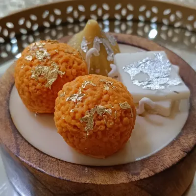 This wooden Indian dessert platter candle is a feast for the eyes. It features handcrafted wax versions of laddoos, a modak, and kaju katli, complete with gold and silver leaf details, all set in a beautiful wooden bowl.