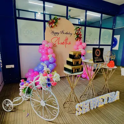 A wide view of the birthday setup for 'Shwetha', featuring a personalized arched backdrop, pink and purple balloon garlands, and a decorative white bicycle prop with flowers.
