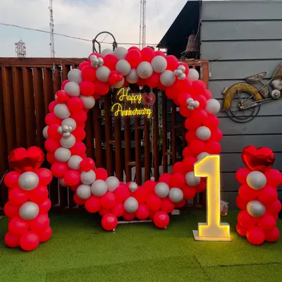 Celebrate your first year together with this vibrant outdoor setup. A circular balloon arch in red and silver frames a neon "Happy Anniversary" sign, with a large light-up number "1" making it a perfect photo opportunity.