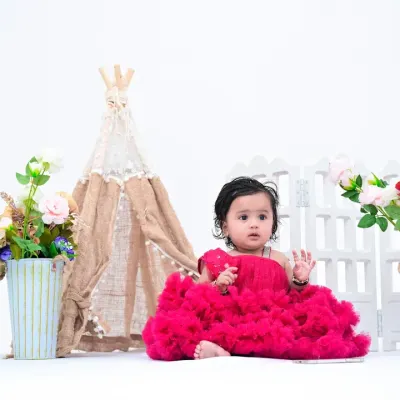 A beautiful baby girl in a vibrant pink dress sits against a clean, white background. This simple setup, with just a few floral and teepee props, keeps the focus entirely on her.