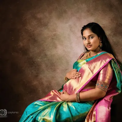 A beautiful, painterly-style maternity portrait of a woman seated in a stunning silk saree. The warm, textured backdrop and soft lighting give the photograph a timeless, classic painting feel.