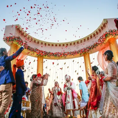 A grand varmala moment under a beautiful arch, with guests showering the couple with rose petals. This wide shot captures the scale and joy of the celebration as the couple officially begins their life together.