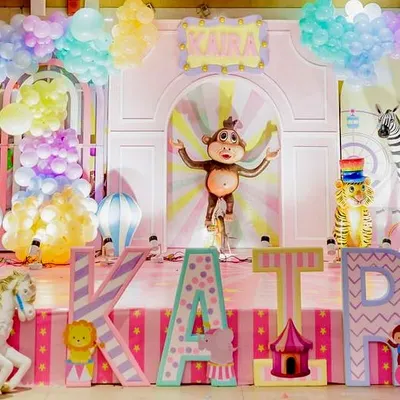 A vibrant, pastel colored carnival stage with a monkey on a unicycle as the centerpiece. Large, custom letters spelling the birthday girl's name are decorated with different patterns and characters.