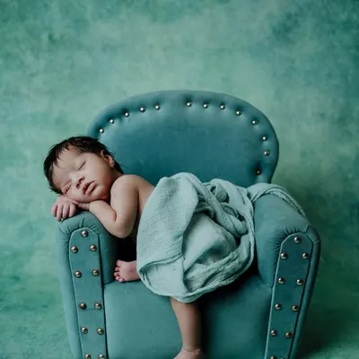 I create calm and artistic settings for newborn photoshoots. Here, a sleeping baby is posed on a miniature teal armchair, with a matching backdrop and soft wrap, highlighting the peaceful and styled nature of my newborn art sessions.