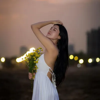 A beautiful beach portrait of a woman in a white dress holding yellow flowers, with the city lights creating a soft bokeh in the background during twilight.