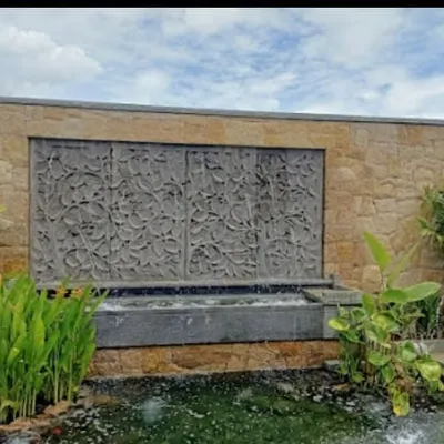 A large, ornate water feature with a carved stone panel, set against a natural stone wall. Water cascades into a basin surrounded by aquatic plants, creating a stunning focal point.