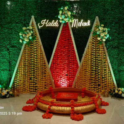 A unique Haldi and Mehendi backdrop featuring a green foliage wall and triangular structures decorated with marigold and jasmine garlands. A neon sign adds a modern touch.