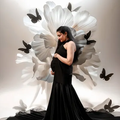 A masterpiece of elegance. The mom-to-be in a stunning black gown is framed by a giant white paper flower and delicate black butterflies, creating a high-fashion, artistic portrait.