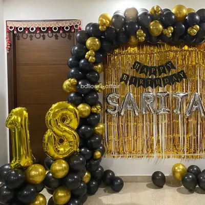 A glamorous 18th birthday decoration in black and gold. This home setup includes a balloon arch, a gold shimmer backdrop, and large foil number balloons.