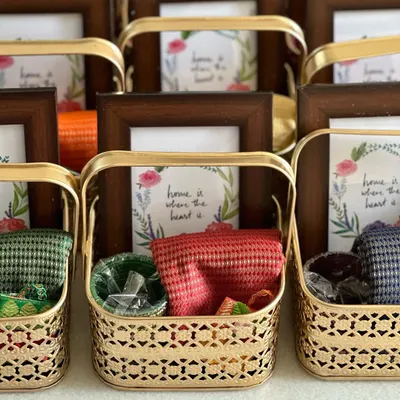 A beautiful housewarming return gift set. Each golden metal basket contains a custom photo frame, a colorful blouse piece, and other traditional items, making it a thoughtful and complete favor.