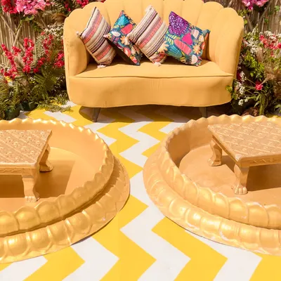 A picture-perfect Haldi seating area. This setup features a bright yellow sofa against a stunning backdrop of a bamboo lattice and a vibrant red bougainvillea arch.