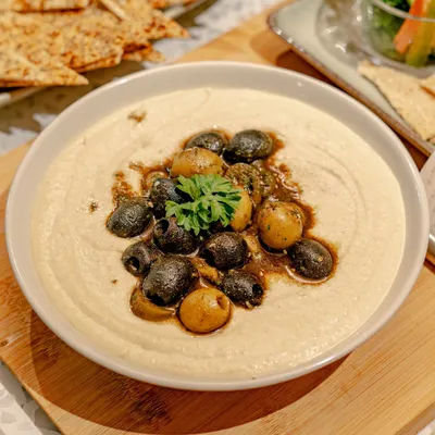 A close-up of our classic hummus, topped with olives and a drizzle of olive oil. Served on a wooden board, it's a simple yet elegant addition to any grazing table.