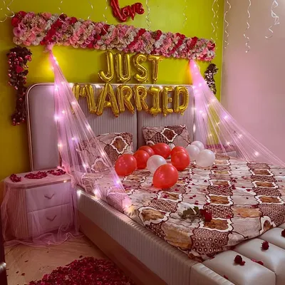 A classic 'Just Married' room decoration to welcome the newlyweds. This setup includes foil balloons, a floral arrangement on the headboard, a canopy with fairy lights, and a floor covered in rose petals.