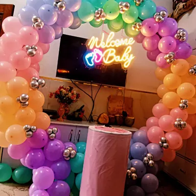 A vibrant and colorful welcome home decoration featuring a circular rainbow balloon arch. The "Welcome Baby" neon sign adds a modern and celebratory touch to the setup.