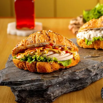 A hearty croissant sandwich filled with fresh ingredients. The rustic stone platter and warm lighting emphasize the sandwich's gourmet quality and the flakiness of the multigrain croissant.