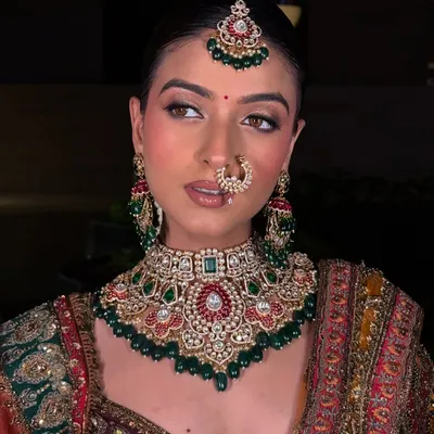A perfect portrait of a modern bride wearing a traditional multi-gemstone choker. This Polki and Kundan set features rich green and red stones, complete with a matching nath and maang tikka for a regal look.