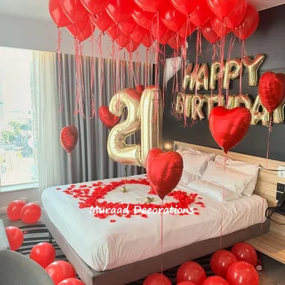 A classic romantic birthday surprise. This room is decorated with red ceiling balloons, 'Happy Birthday' foil letters, large number balloons, and a heart made of rose petals on the bed.