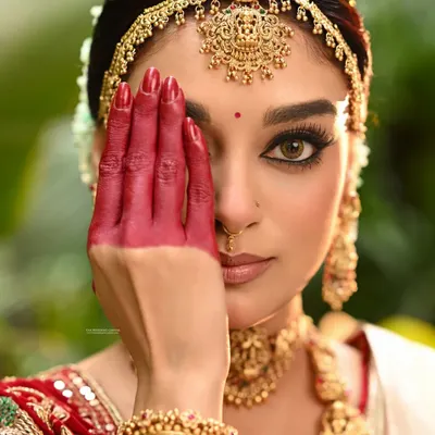 A powerful bridal close-up where the model's hand, decorated with red 'alta', partially covers her face. This creative composition draws attention to her intense eyes and the traditional elements of her look.
