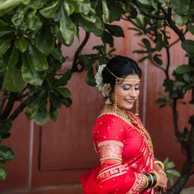 A timeless portrait of a Maharashtrian bride. We captured her gentle, downward glance and smile, highlighting the traditional jewelry, the mundavalya on her forehead, and the vibrant red of her wedding saree against a natural green background.