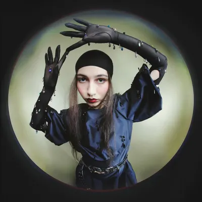 A distorted fisheye portrait from the 'Peek-a-boo' series. The model, wearing embellished gloves and a head covering, frames her face in a playful yet unsettling pose, creating a surreal, peephole effect.