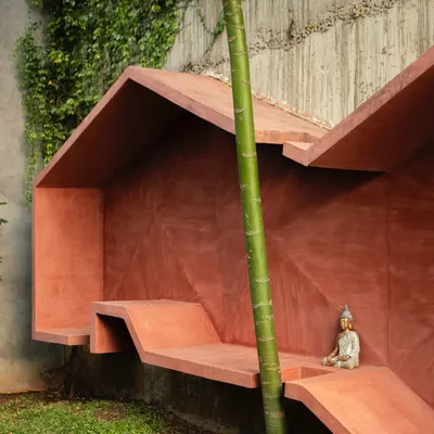 A bold, monolithic seating structure in a striking red oxide finish. Tucked against a vine-covered wall, this piece blurs the line between sculpture and functional bench, creating a quiet moment of stillness in the garden.