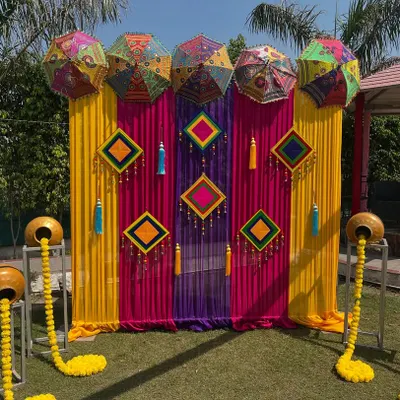 A vibrant photo booth I created for a Mehndi function. It features a backdrop of colorful drapes, Rajasthani umbrellas, and traditional hangings, with decorated pots adding a rustic touch.