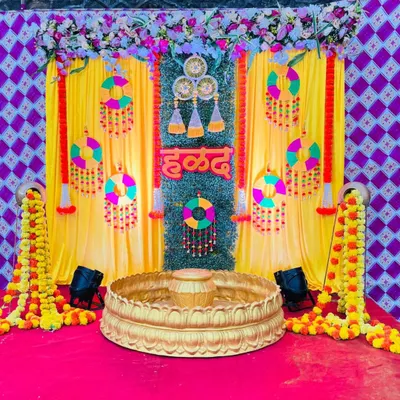 A vibrant Haldi ceremony backdrop featuring bright yellow curtains and a central panel of artificial grass. The setup is decorated with colorful hanging ornaments, marigold garlands, and a 'हळद' sign, creating a festive atmosphere.