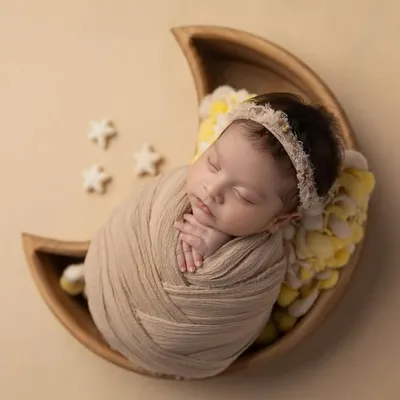 A close up of a newborn swaddled in a neutral wrap, resting in a wooden moon bowl. The yellow floral details and tiny stars add a touch of whimsy to this dreamy setup.