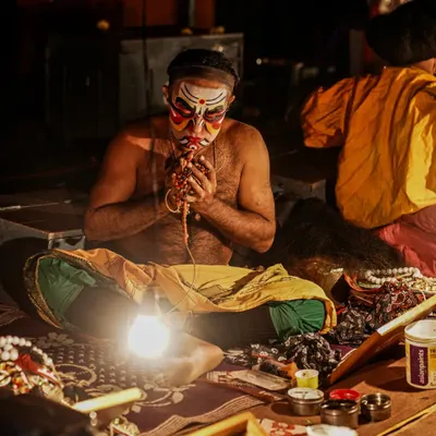 An intimate look at a Yakshagana artist in the 'Chowki' as he meticulously applies his makeup. This close-up shot captures the intense focus and ritualistic nature of the transformation process before the performance begins.