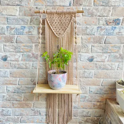 This single-tier hanging shelf features a V-shaped macrame panel with long fringe, crafted in a neutral beige cord. It’s a versatile design that fits beautifully in rustic, boho, or minimalist spaces, perfect for displaying a small potted plant.